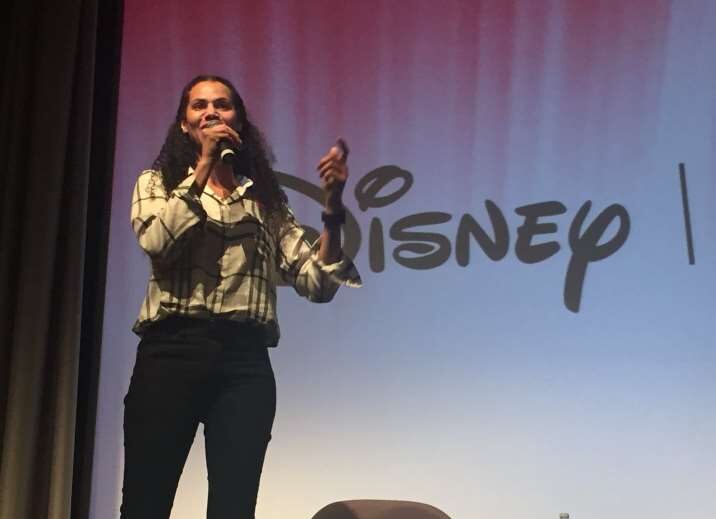 A women with black hair, a white shirt, and black pants holds a microphone and speaks on a stage in front of the Disney logo