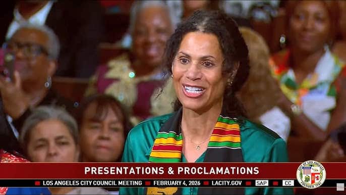 A woman with black hair, wearing a green blouse and a red, yellow, green, and black striped stole, speaks on camera.