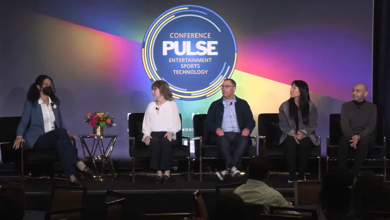 A woman wearing a black surgical mask is seated to the far left of panelists on a stage in front of a sign that reads 