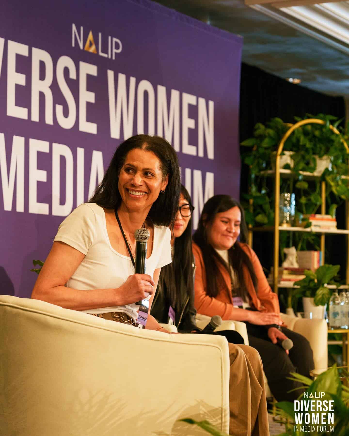 A women with black hair and a white shirt is seated to the right of two other panelists,. She holds a microphone and looks over her shoulder and smiles