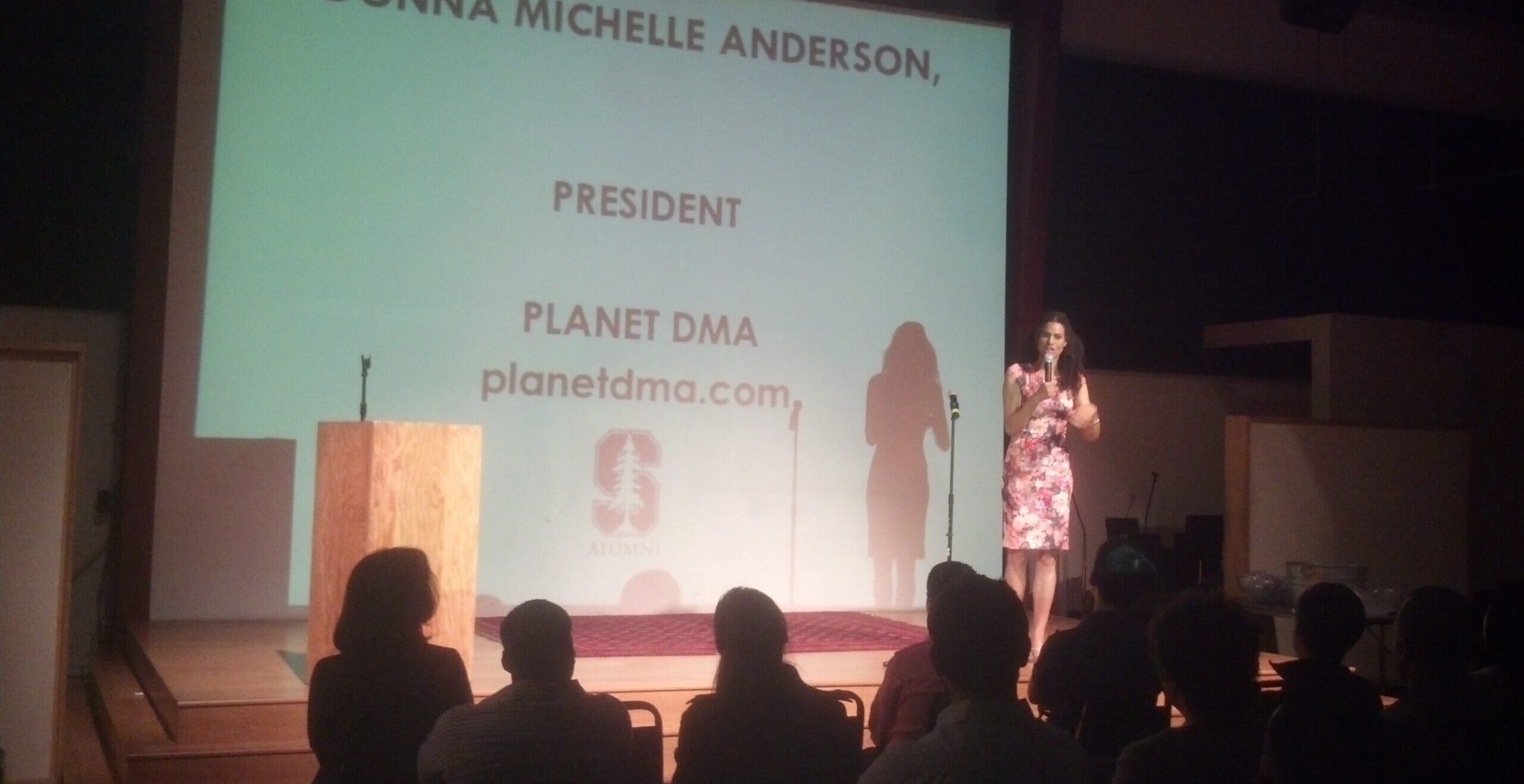 A women with black hair and a floral dress stands on stage in front of a sign that says "President, Planet DMA" above the Stanford logo.